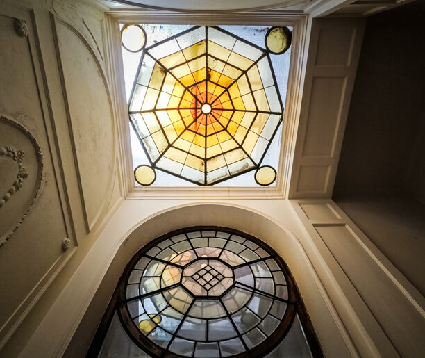 Fotografia przedstawia sufit wnętrza budynku przy ulicy Karmelickiej 45. Sufit jest ozdobiony kolorowym witrażem i dekoracyjnymi sztukateriami.