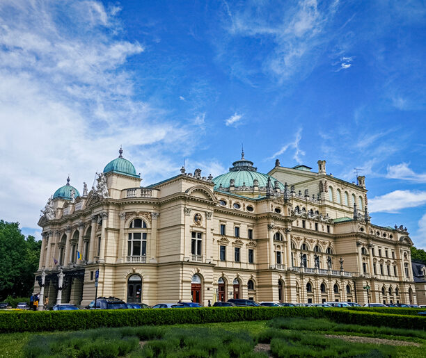 Zdjęcie przedstawia widok na klasyczny budynek teatru z kopułami na dachu i bogato zdobioną fasadą. W tle widać niebieskie niebo z delikatnymi chmurami.
