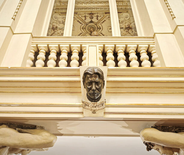 Zdjęcie przedstawia wnętrze teatru, pokazując ozdobny balkon i popiersie Jana Zawieyskiego. Wnętrze jest eleganckie, z bogatymi zdobieniami i jasnym wystrojem.