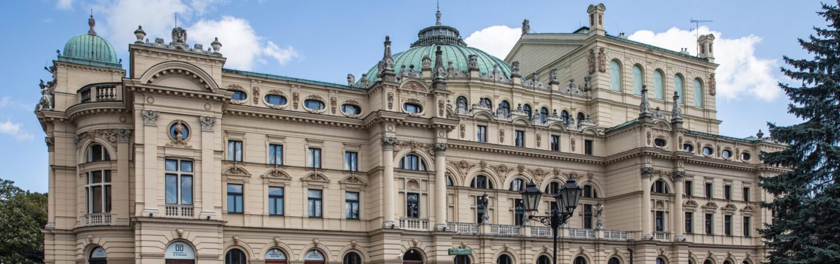 Zdjęcie przedstawia widok na klasyczny budynek teatru z kopułami na dachu i bogato zdobioną fasadą. W tle widać niebieskie niebo z delikatnymi chmurami.