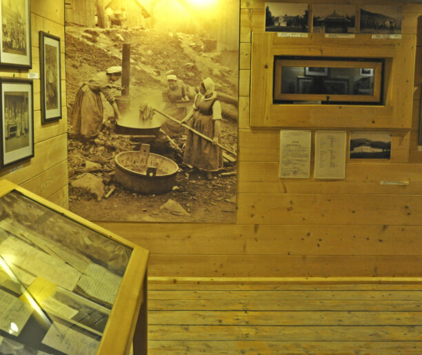 Ekspozycja muzealna z wielkim zdjęciem prac kuchennych: Wnętrze muzeum z wystawą starych fotografii na drewnianej ścianie, w tym dużego zdjęcia przedstawiającego kobiety pracujące przy gotowaniu na zewnątrz. Obok znajdują się gabloty z dokumentami.