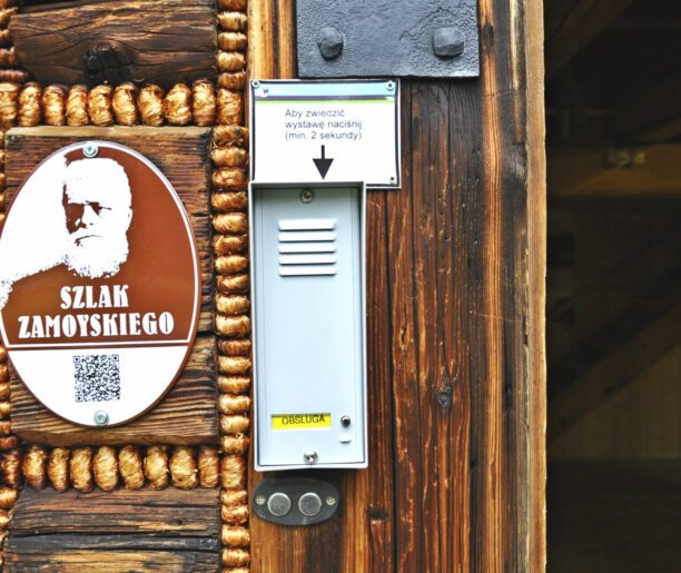 Tabliczka Szlaku Zamoyskiego przy interkomie: Fotografia tabliczki Szlaku Zamoyskiego umieszczonej na drewnianej ścianie obok interkomu. Tabliczka zawiera wizerunek Władysława Zamoyskiego i kod QR, a nad interkomem znajduje się instrukcja jego obsługi.