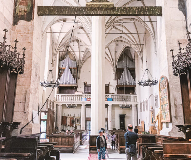 Wnętrze zabytkowego kościoła z widokiem na organy, wysokie sklepienia i ludzi zwiedzających. Drewniane ławki i dekoracyjne żyrandole.