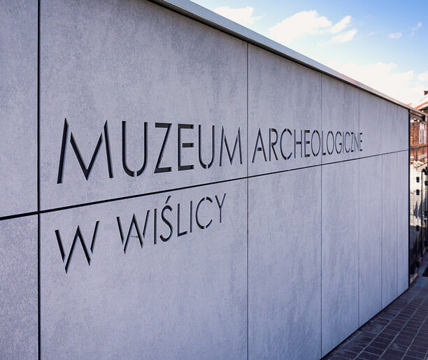 Zewnętrzna ściana Muzeum Archeologicznego w Wiślicy z napisem nazwy instytucji na tle nieba.