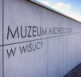 Zewnętrzna ściana Muzeum Archeologicznego w Wiślicy z napisem nazwy instytucji na tle nieba.