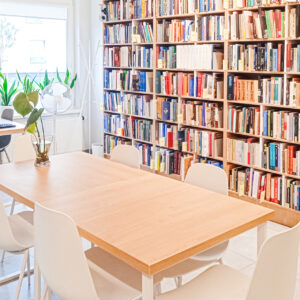 Jasne pomieszczenie z dużym stołem i białymi krzesłami w otoczeniu regałów pełnych książek. Rośliny doniczkowe dodają wnętrzu świeżości i przytulności