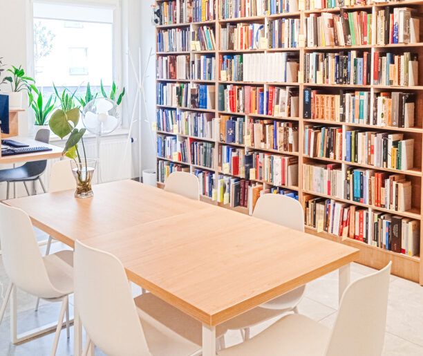 Jasne pomieszczenie z dużym stołem i białymi krzesłami w otoczeniu regałów pełnych książek. Rośliny doniczkowe dodają wnętrzu świeżości i przytulności