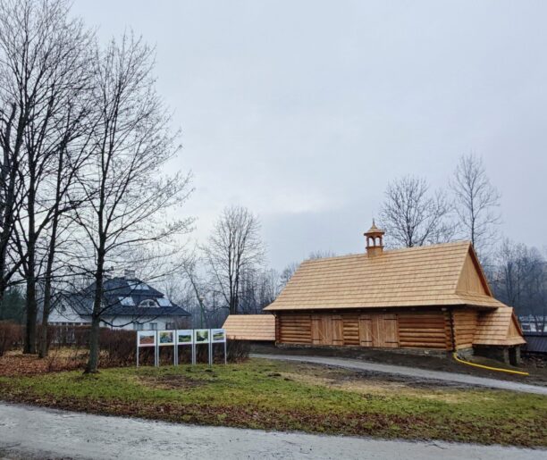 Drewniany budynek wozowni w stylu regionalnym z nowym dachem krytym gontem, otoczony trawą i bezlistnymi drzewami.