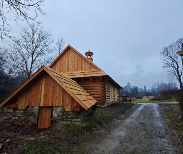 Drewniana wozownia widziana z boku, z widocznymi detalami gontowego dachu i kamiennego fundamentu, prowadzi do niej żwirowa ścieżka.