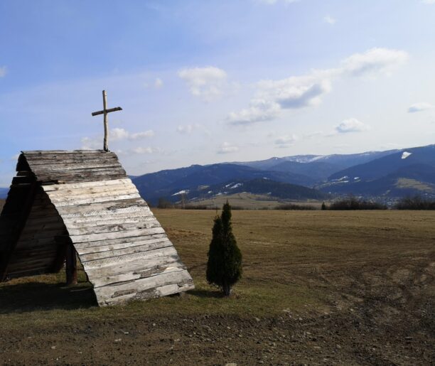 Drewniana kapliczka w kształcie trójkąta z krzyżem na szczycie, stojąca na otwartym polu z widokiem na pasmo górskie w oddali.
