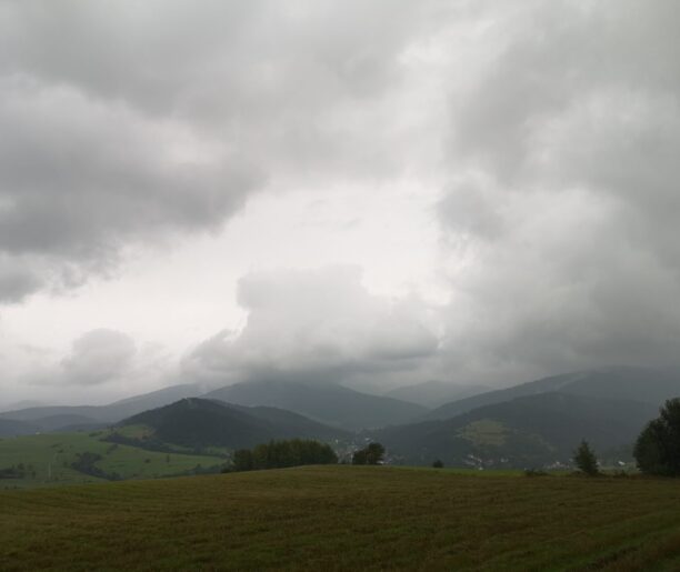 Panorama górska w pochmurny dzień, z widocznymi wzniesieniami, polami i nisko zawieszonymi chmurami.