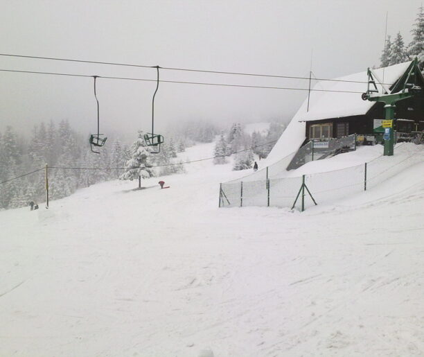 Stacja kolejki linowej w gęstej mgle, z ośnieżonym budynkiem i pustymi krzesełkami na linie.