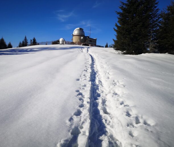 Kopuła obserwatorium astronomicznego na śnieżnym wzgórzu, z wydeptaną ścieżką prowadzącą na szczyt i niebieskim niebem w tle.