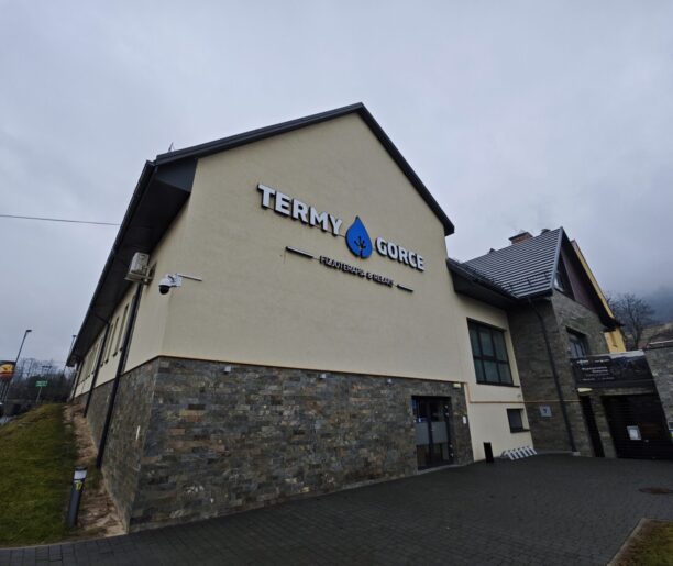 Nowoczesny budynek Term Gorce z logo na fasadzie, otoczony brukowanym chodnikiem i trawnikami, w pochmurny dzień.